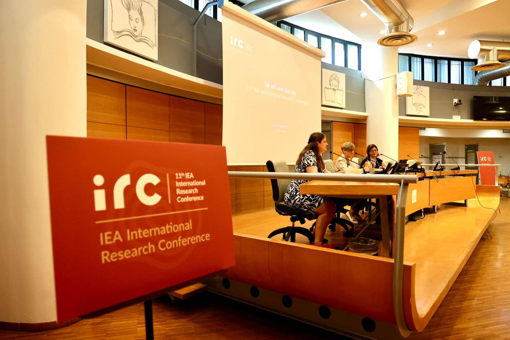 Image of a stage, three speakers in a warmly lit auditorium 